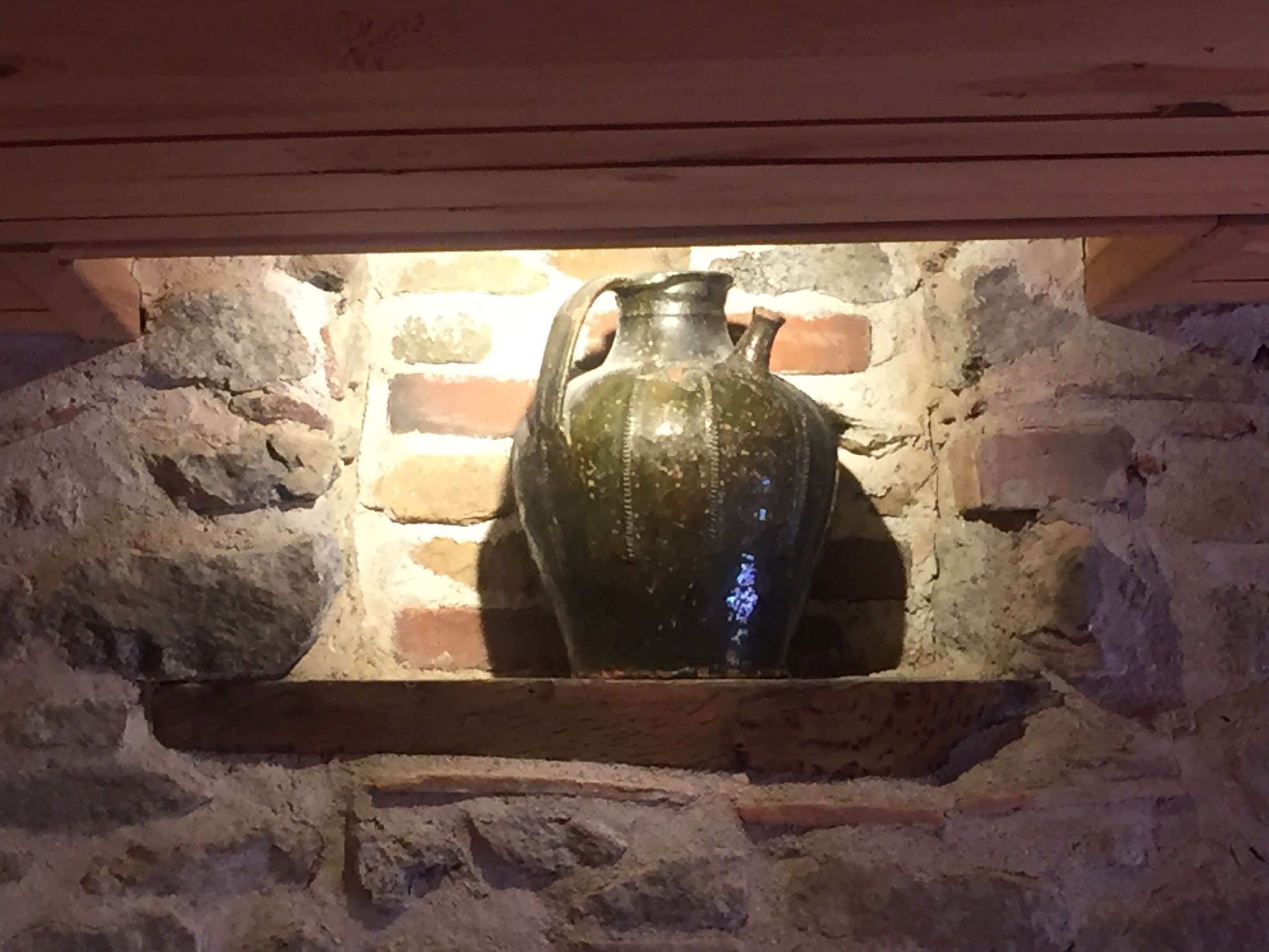 A stone wall with a wooden shelf holding a large, dark-colored ceramic jug.