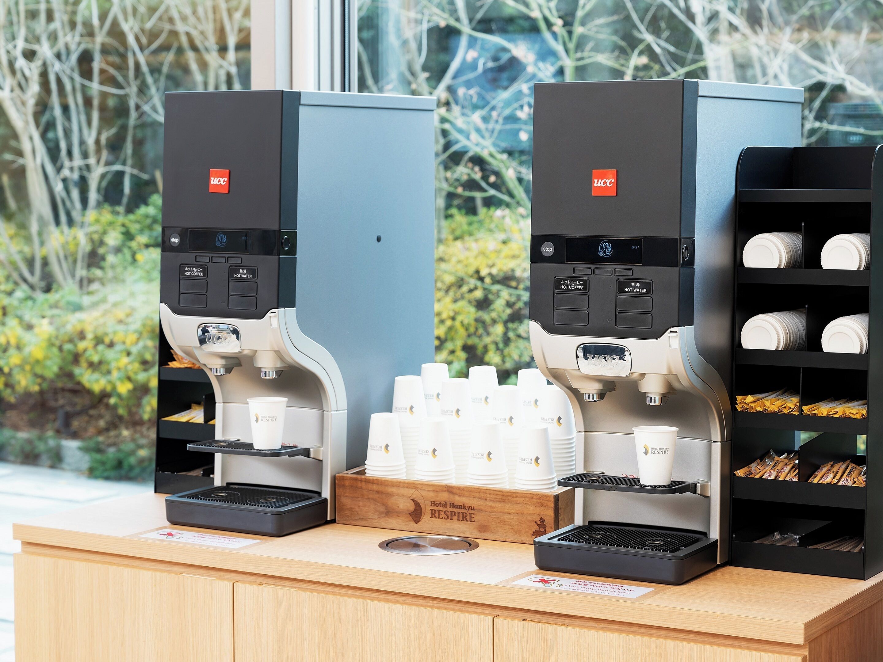 Two coffee machines with hot coffee and hot water options, a wooden counter with a sign reading "Hotel Hankyu RESPIRE", and a shelf with plates and napkins.