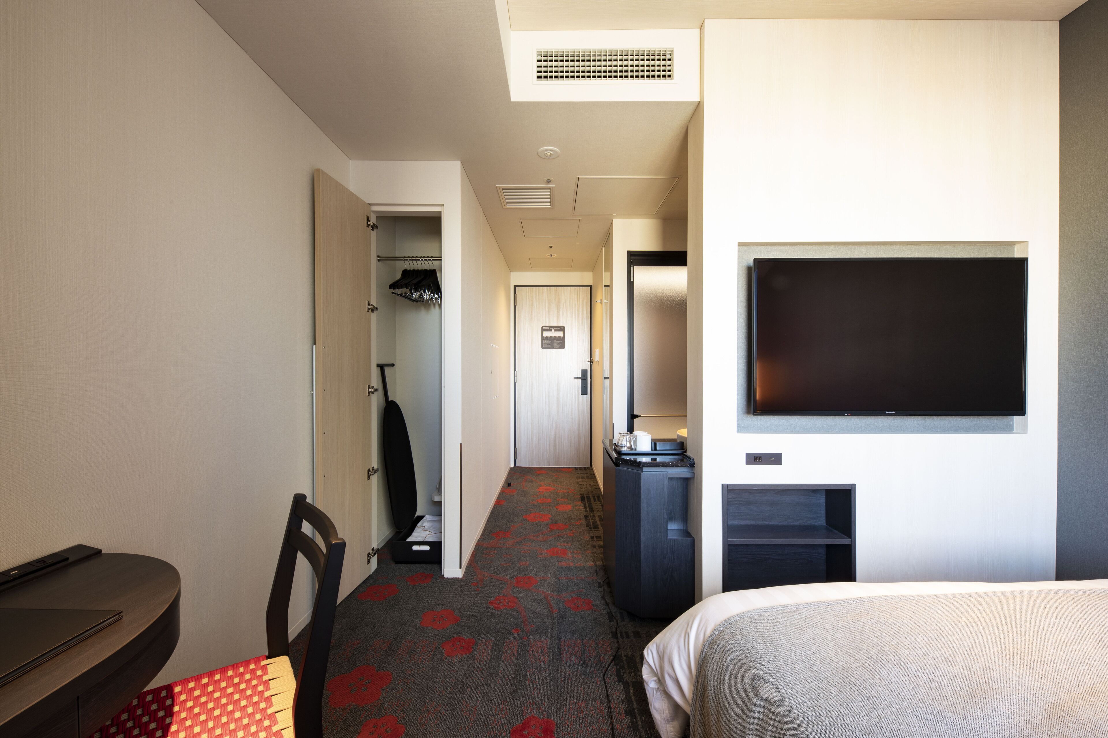 A hotel room with a bed, a desk, a chair, a TV, a wardrobe, and a carpet with a red pattern.