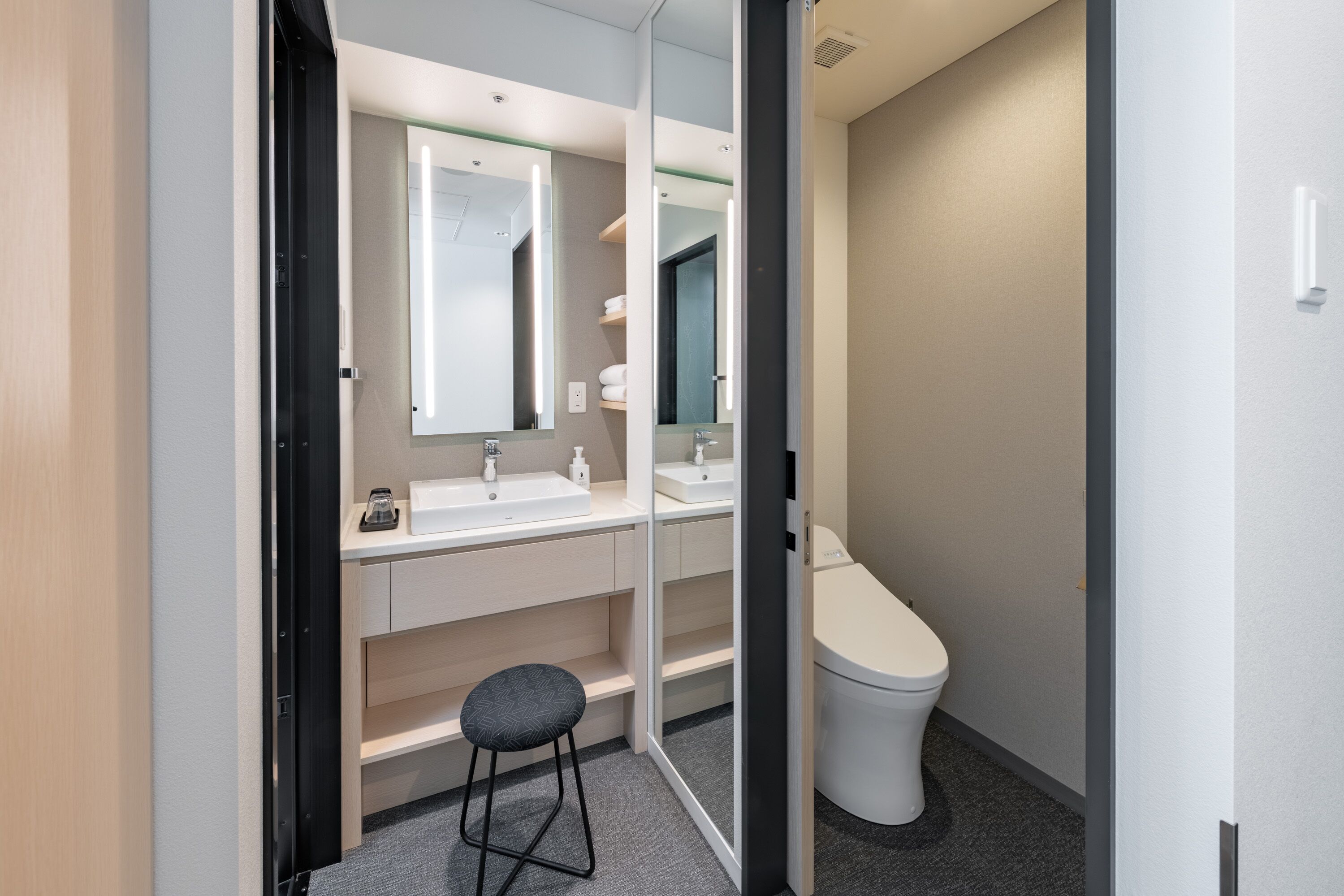 A modern bathroom with a white sink, mirror, and toilet. There is a stool and a shelf with toiletries.