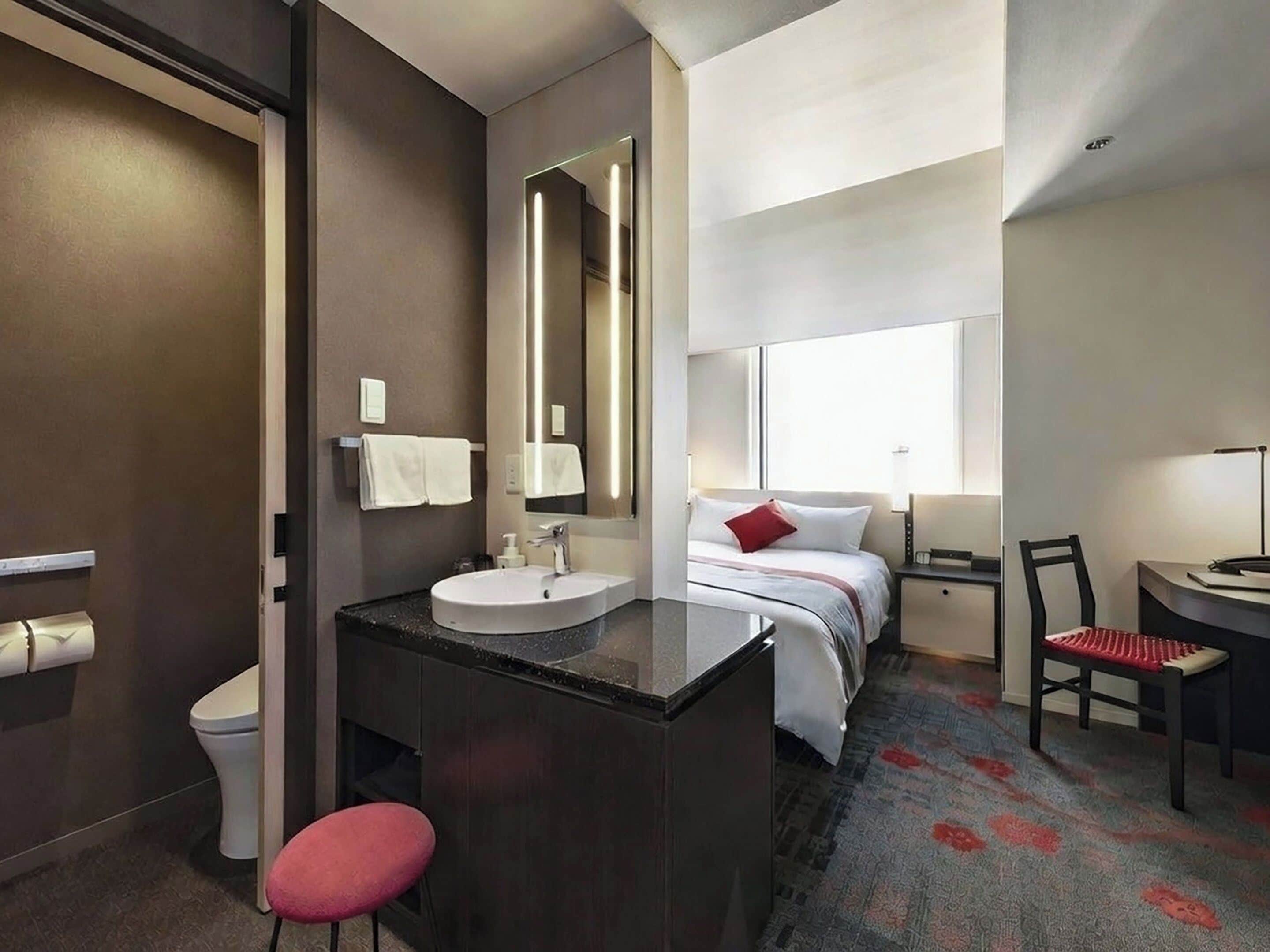A modern hotel room with a bathroom, a bed with a red pillow, a desk with a chair, and a large mirror.