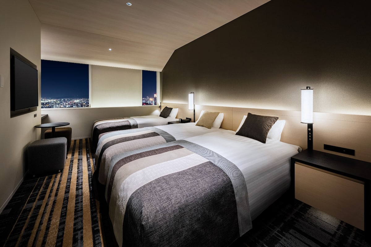 Ein Hotelzimmer mit zwei Betten, einem Fernseher, einem Schreibtisch und Blick auf die beleuchtete Stadt.