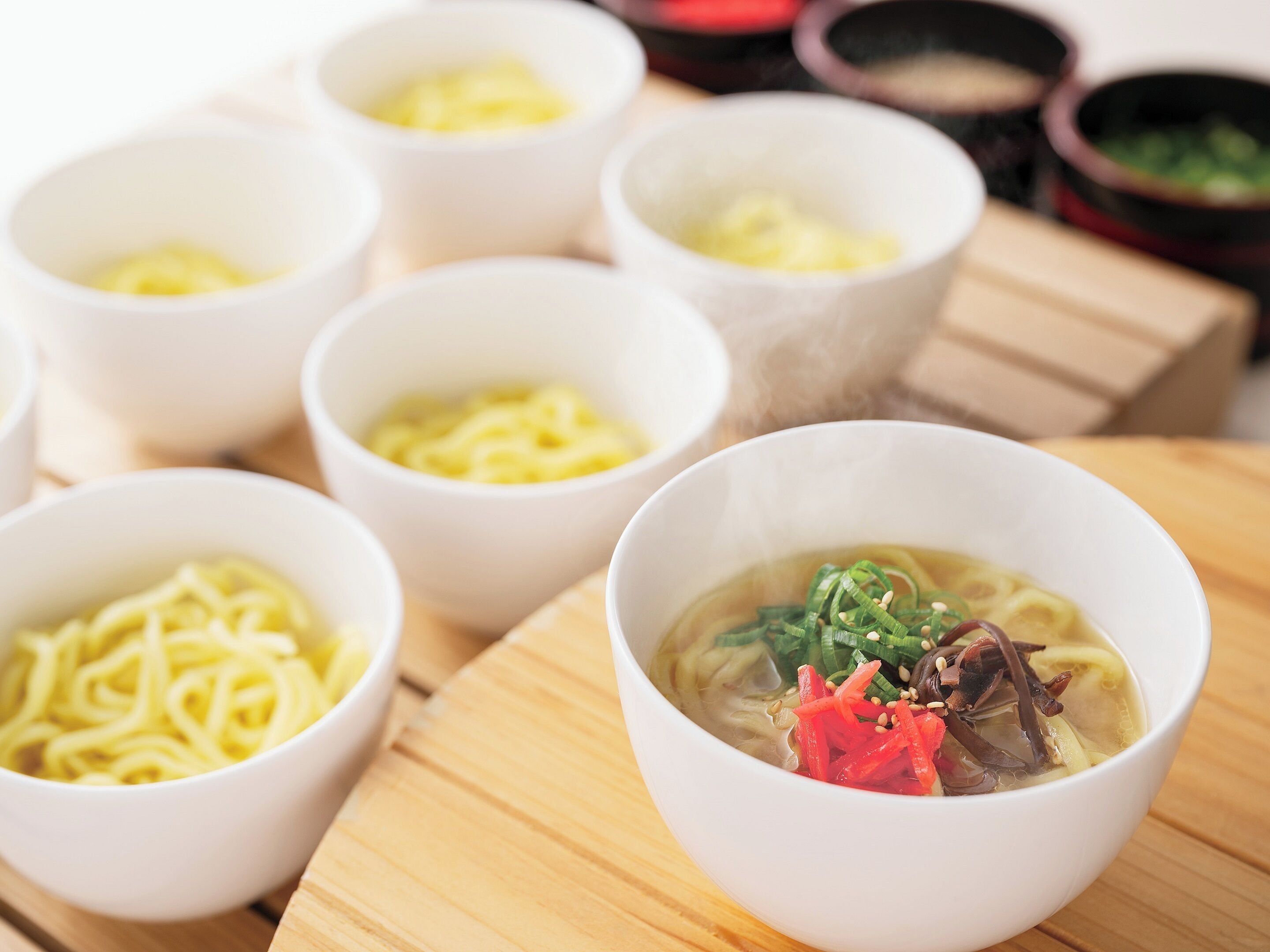 Una mesa con varios cuencos de fideos y sopa, algunos con ingredientes como cebolletas y semillas de sésamo.