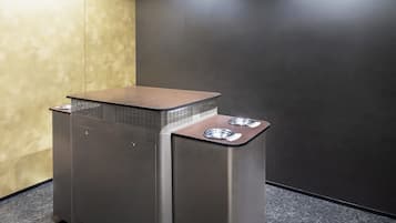 A compact, modern trash bin with a metallic finish and a textured top, placed on a dark floor against a contrasting wall.