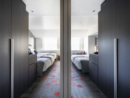Habitación de hotel con armario espejado, dos camas, una mesita y una ventana.