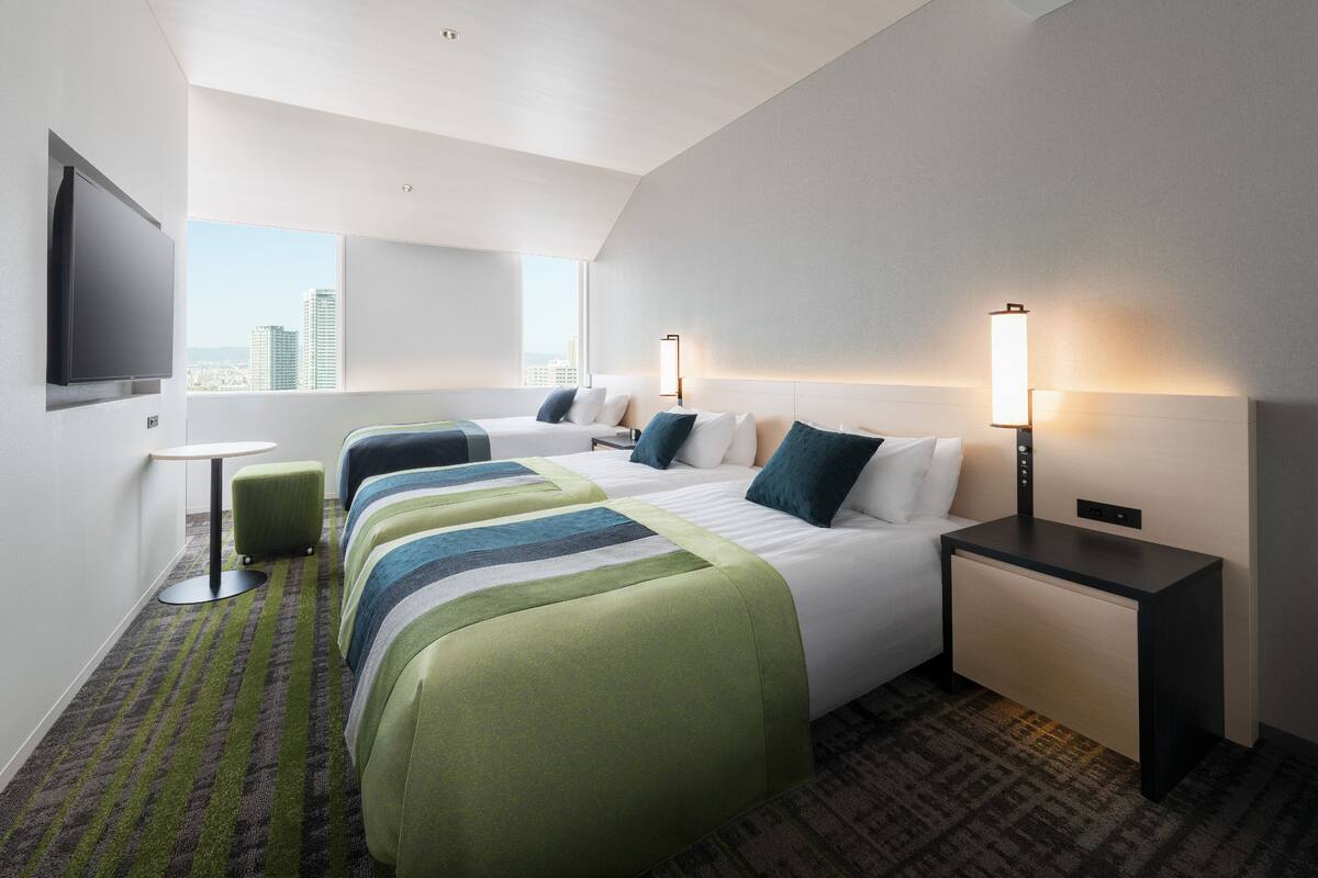 Ein Hotelzimmer mit einem großen Bett, zwei Nachttischen, einem an der Wand montierten Flachbildfernseher und Blick auf die Stadtlandschaft durch das Fenster.