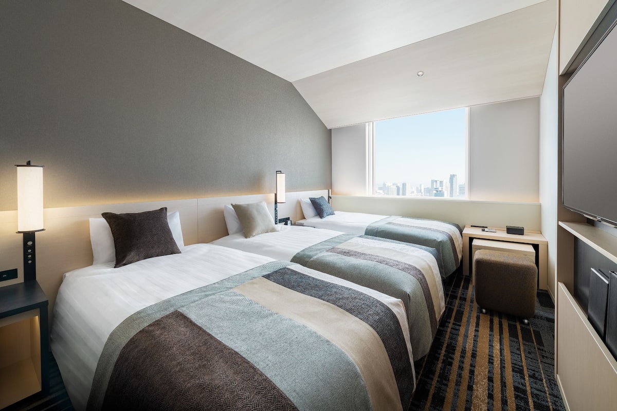Ein Hotelzimmer mit zwei Betten, einem Schreibtisch, einem Stuhl, einem Fernseher und Blick auf die Stadt.