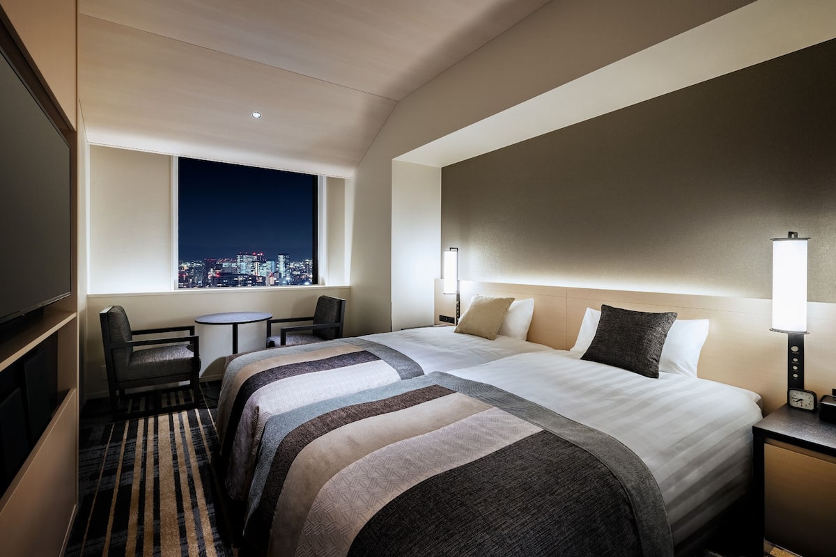 Ein Hotelzimmer mit einem großen Bett, einem Schreibtisch, einem Sessel, einem Fernseher und Blick auf die Stadt.