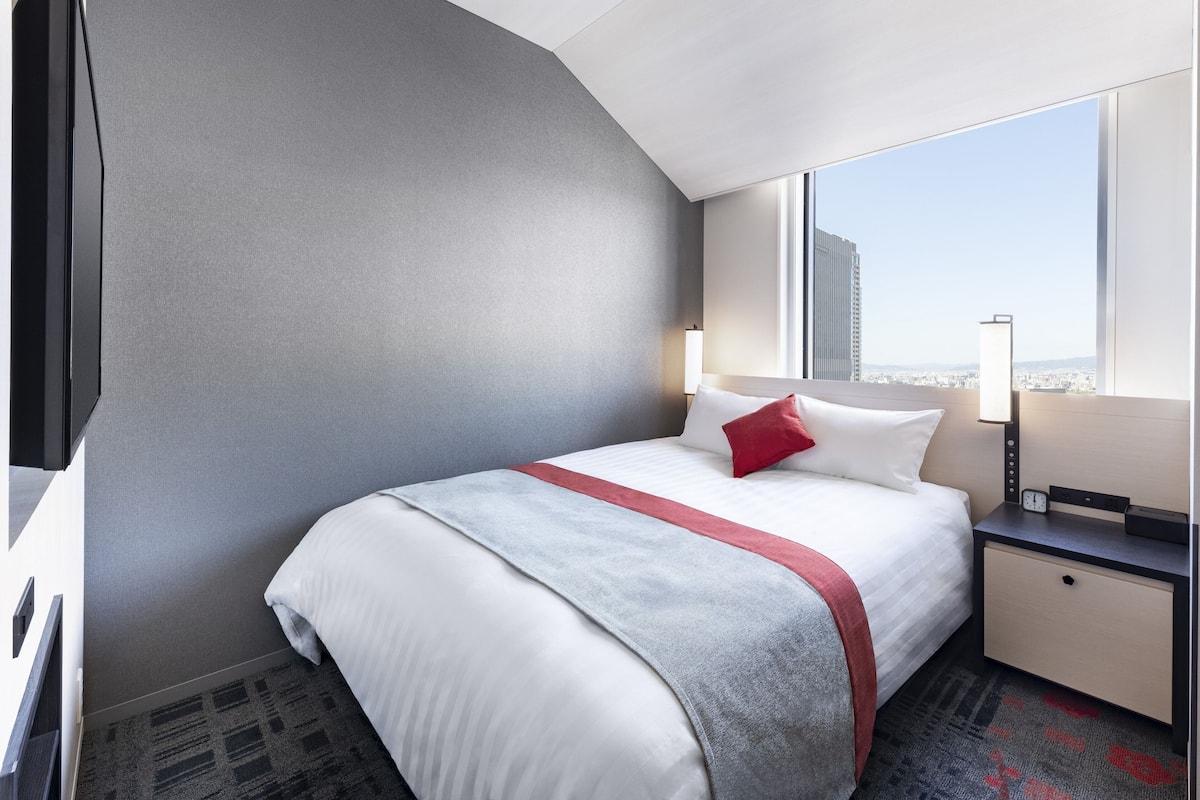 Ein modernes Hotelzimmer mit einem großen Bett, einem Nachttisch, einem Fernseher und Blick auf die Stadt durch das Fenster.