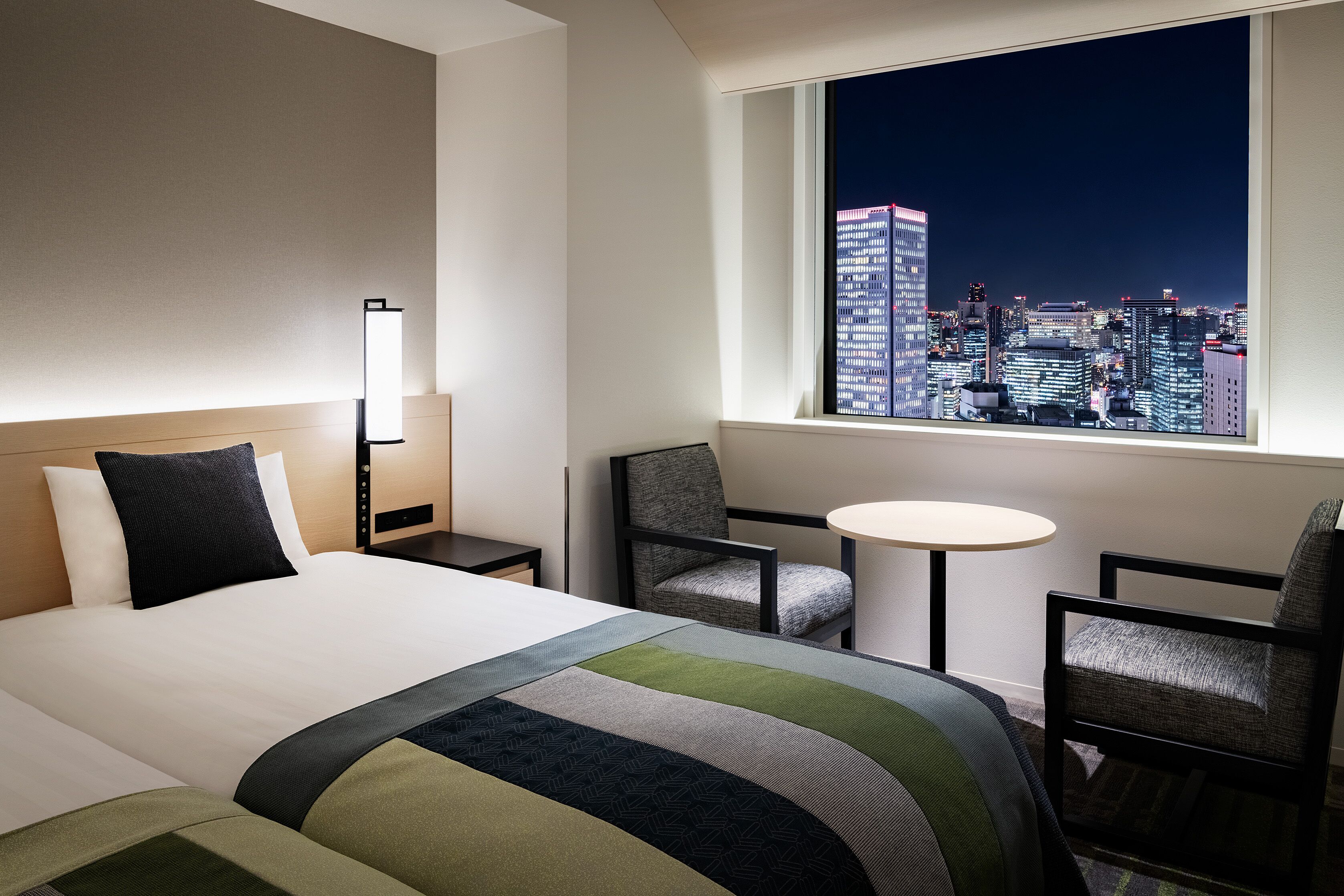 A hotel room with a large bed, two chairs, a small table, and a view of a cityscape at night.