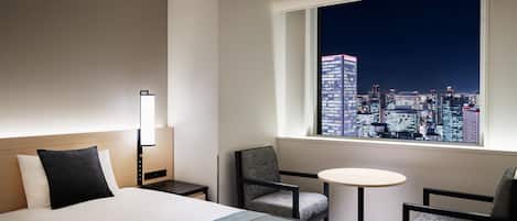 Une chambre d’hôtel avec un grand lit, deux fauteuils, une petite table et une vue sur le paysage urbain illuminé la nuit.