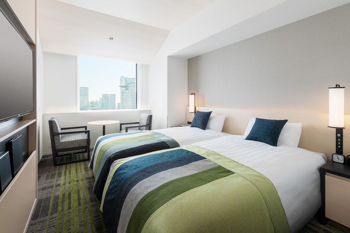 Ein Hotelzimmer mit zwei Betten, einem Fernseher, einem Schreibtisch und einem Fenster mit Blick auf die Stadt.
