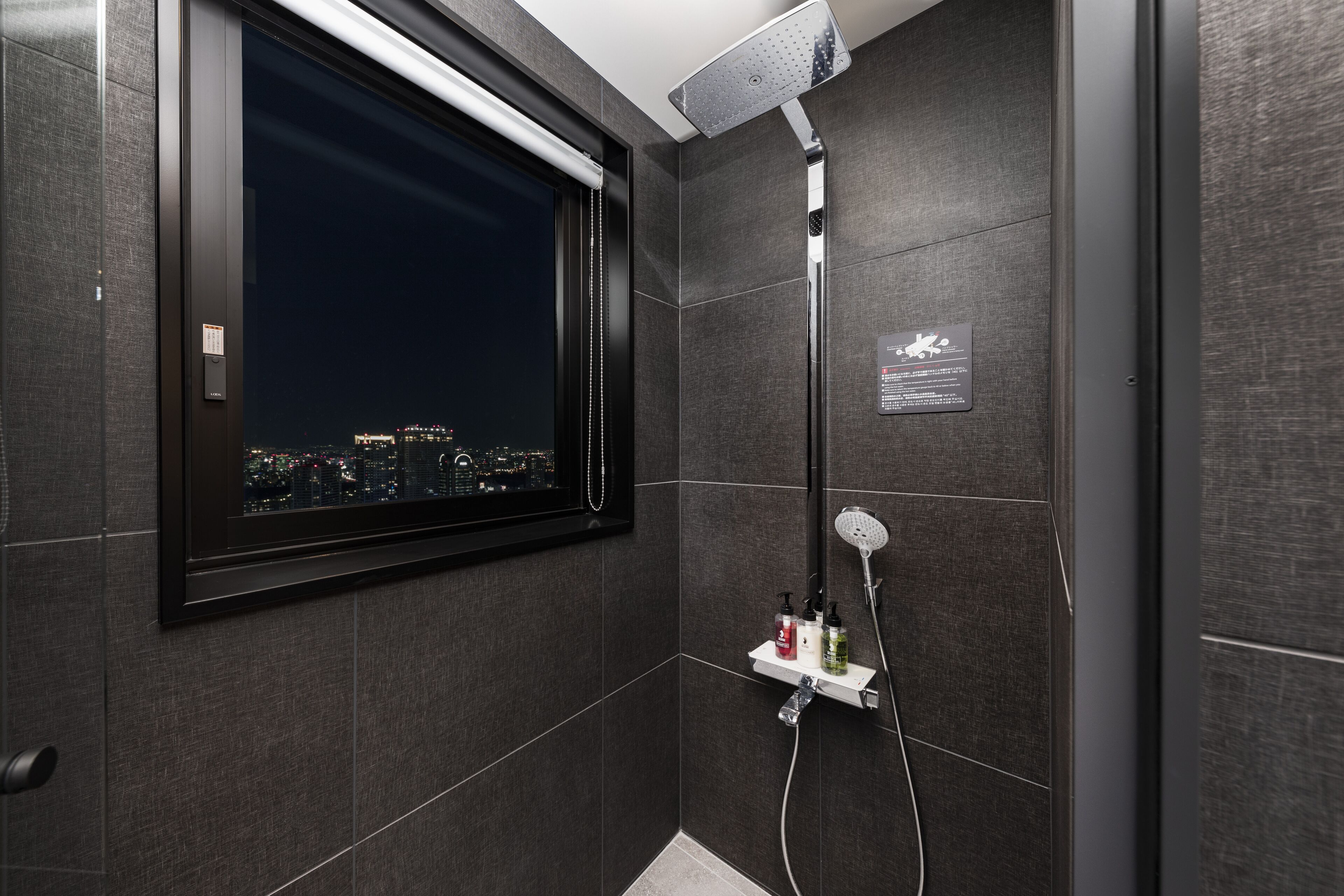 A modern bathroom with a cityscape view through a window, a shower with a handheld head, and a shelf with toiletries.