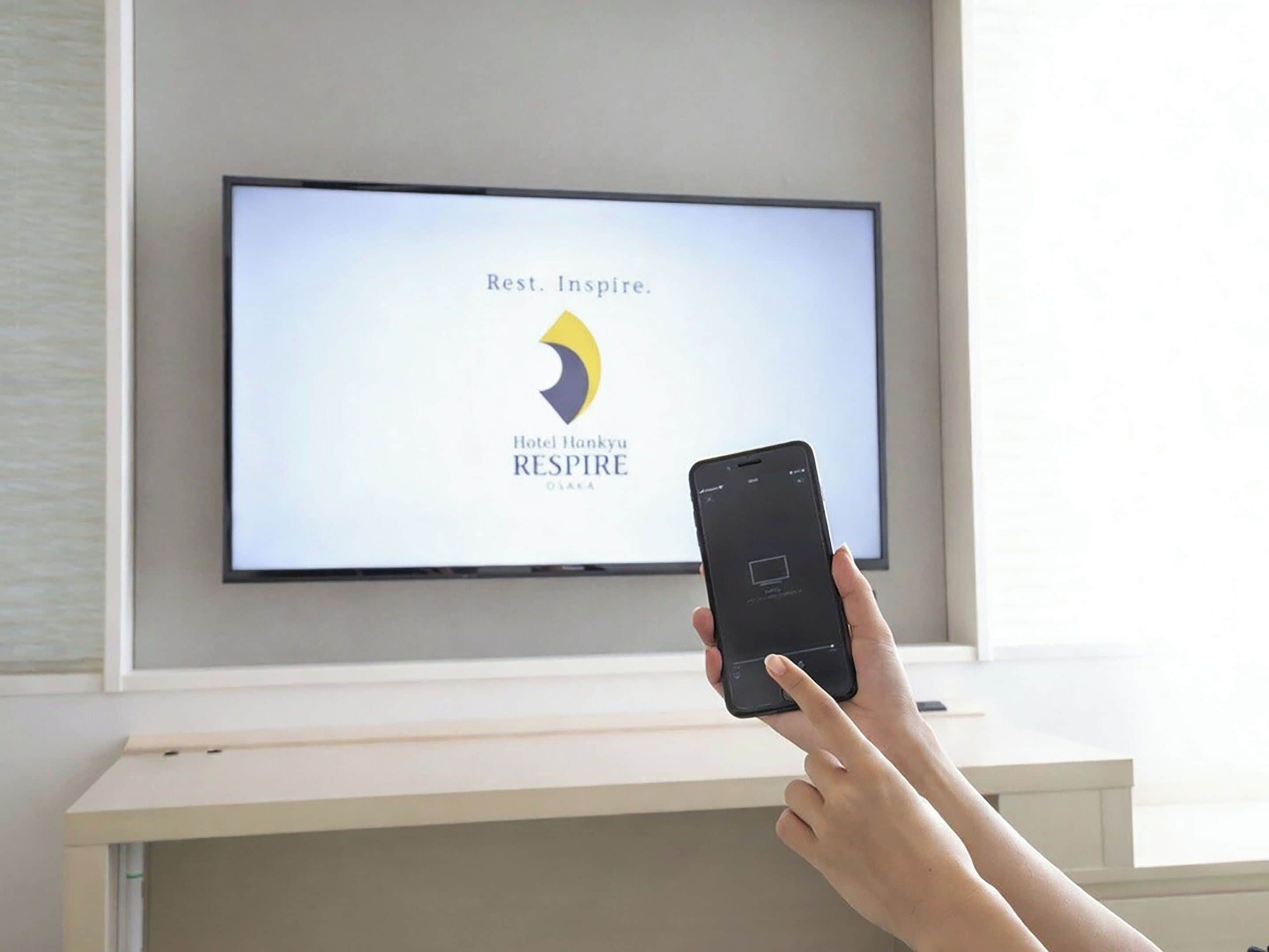 Una persona sosteniendo un teléfono inteligente frente a un televisor de pantalla plana que muestra el logotipo del Hotel Hankyu RESPIRE.