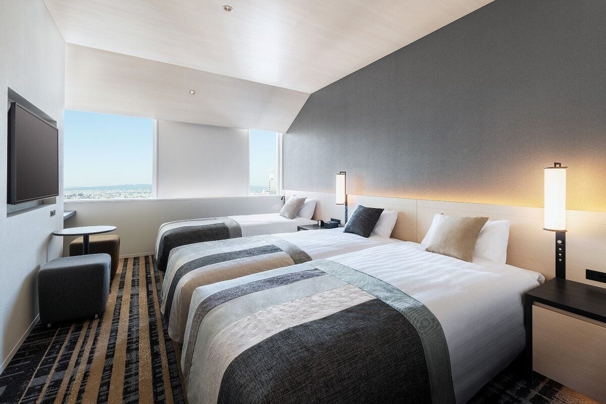 Ein Hotelzimmer mit zwei Betten, einem Schreibtisch, einem Stuhl und einem großen Fenster mit Blick auf die Stadt.
