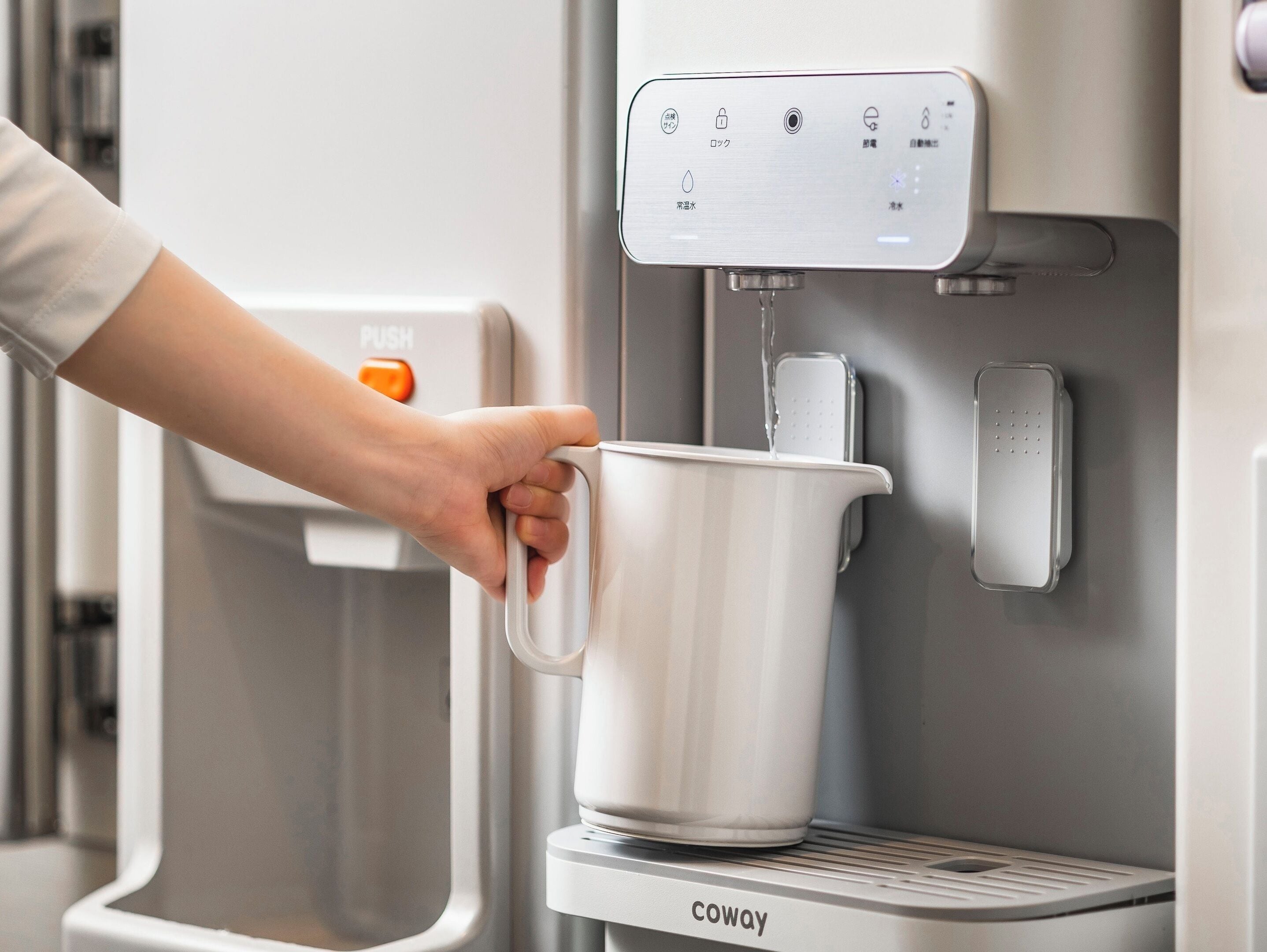Una mano sosteniendo una taza blanca debajo de un dispensador de agua con un panel de control y un botón pulsador.