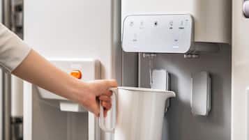 Una mano sosteniendo una taza blanca debajo de un dispensador de agua con un panel de control y un botón pulsador.