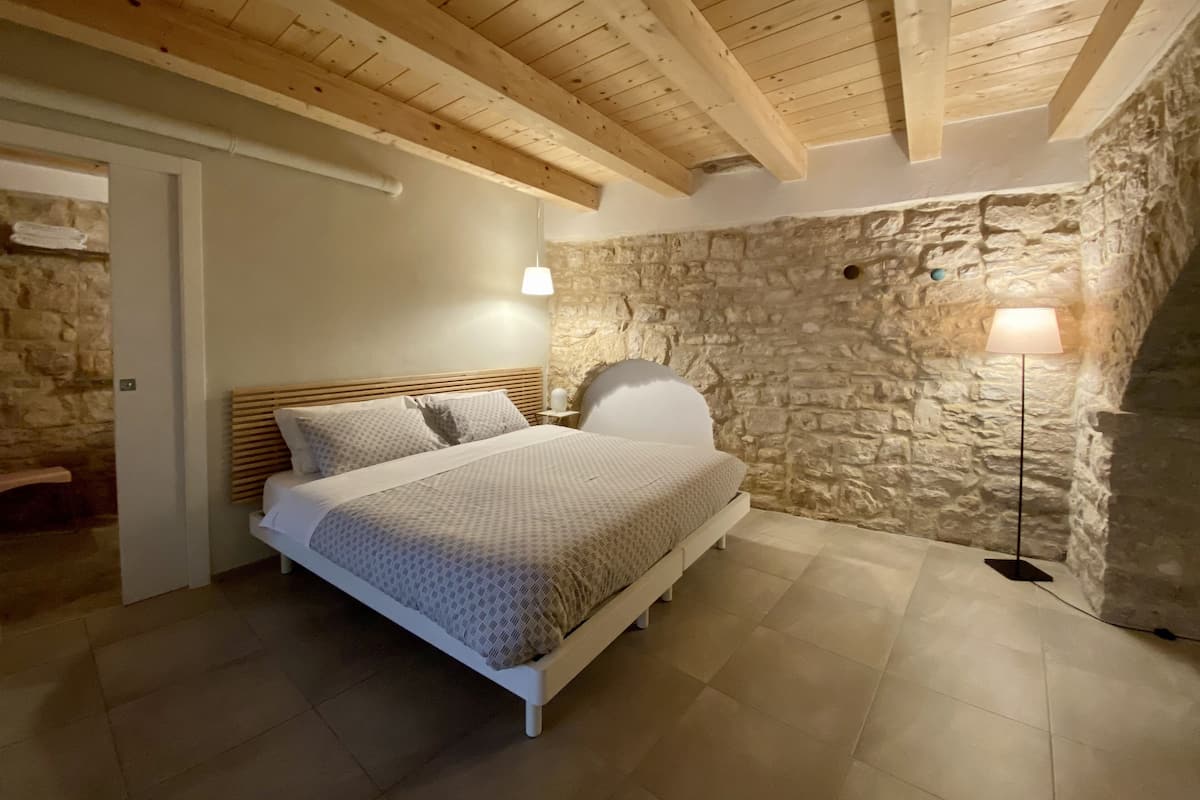 Een slaapkamer met een stenen muur, houten balken in het plafond, een bed met beddengoed met een patroon en een vloerlamp.