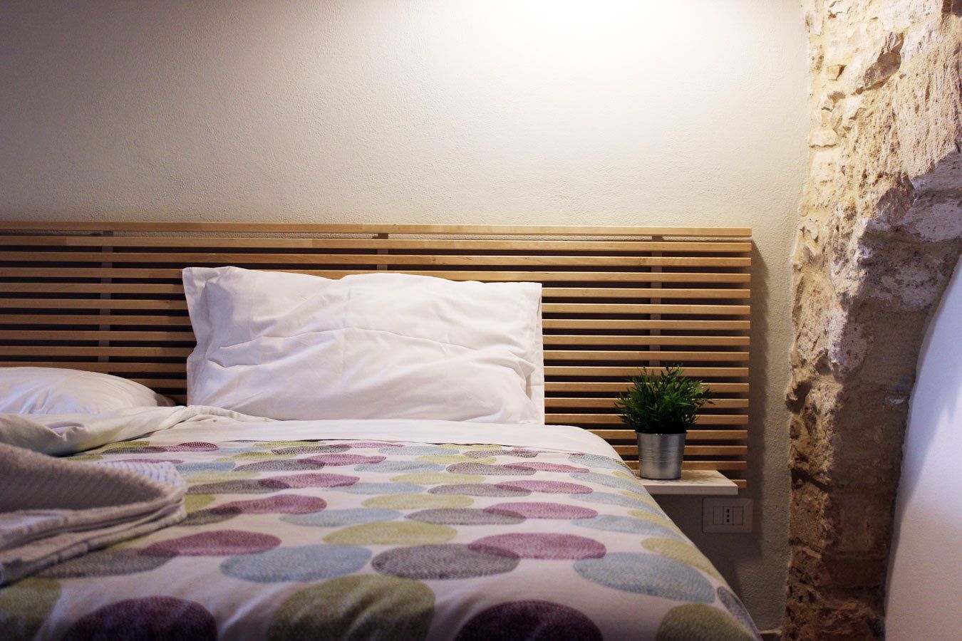 A bed with a wooden headboard, white pillows, and a colorful blanket. There is a small potted plant on the nightstand beside the bed.
