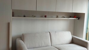 A minimalist living room with a sofa, a built-in shelf with decorative items, and a window. - GuestHost - Our studio is located 5 minutes walk from the beach, in the most beautiful area of Sanremo. With a small terrace with sea view and free unassigned condominium parking, (subject to availability) the apartment is waiting for you in the city of  (Sanremo)