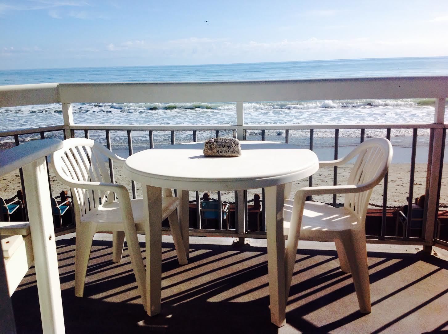 Une table ronde, accompagnée de deux chaises en plastique blanc, est placée sur un balcon donnant sur l’océan.
