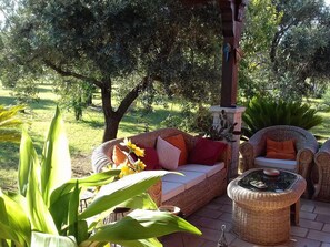 An outdoor seating area with wicker furniture, a glass-top table, and a backdrop of trees and greenery. - B&B Elda Salento (Tuglie)