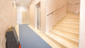 A hallway with a blue carpet, beige walls, a wooden door, and a staircase with beige steps and a metal handrail. - Hotel Mar Garni (Belgrade)