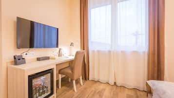 A hotel room with a flat-screen TV mounted on the wall, a desk with a chair, a mini-fridge with beverages, and a large window with curtains.