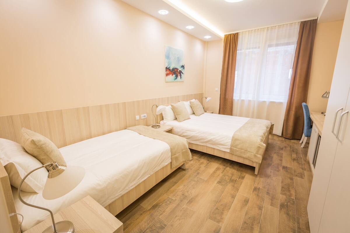 A hotel room with two single beds, a desk, a chair, and a window with curtains.