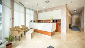 A reception area with a reception desk, several chairs, a potted plant, and a view of the outside through large windows. - Hotel Mar Garni (Belgrade)
