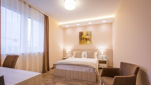 A hotel room with a bed, a desk, and a chair. There is a painting on the wall and a window with curtains. - Hotel Mar Garni (Belgrade)