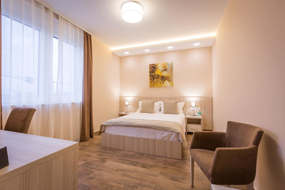 A hotel room with a bed, a desk, and a chair. There is a painting on the wall and a window with curtains.