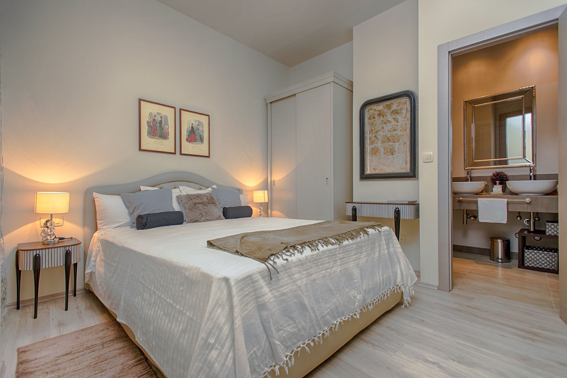 A modern bedroom with a large bed, bedside tables, a nightstand, a bathroom with a sink and mirror, and framed artwork on the walls.