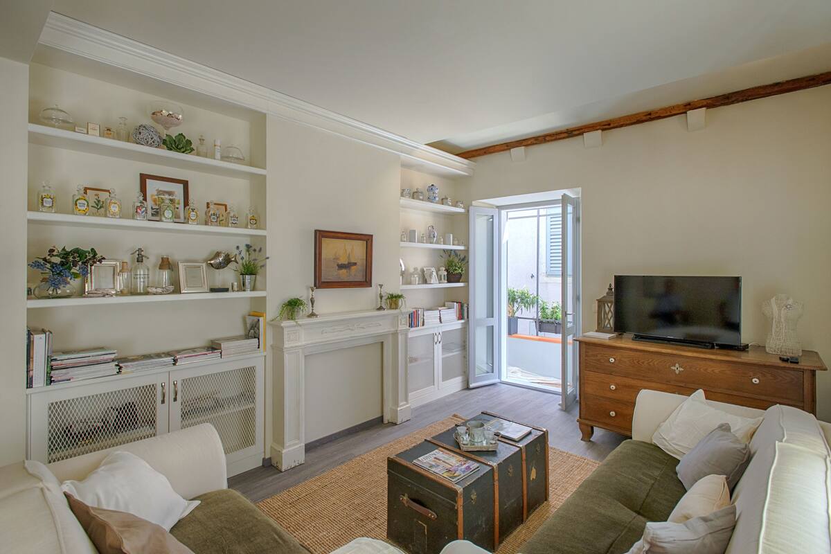 Een woonkamer met een bank, salontafel en televisie. Er staan planken met decoratieve objecten en een open haard.