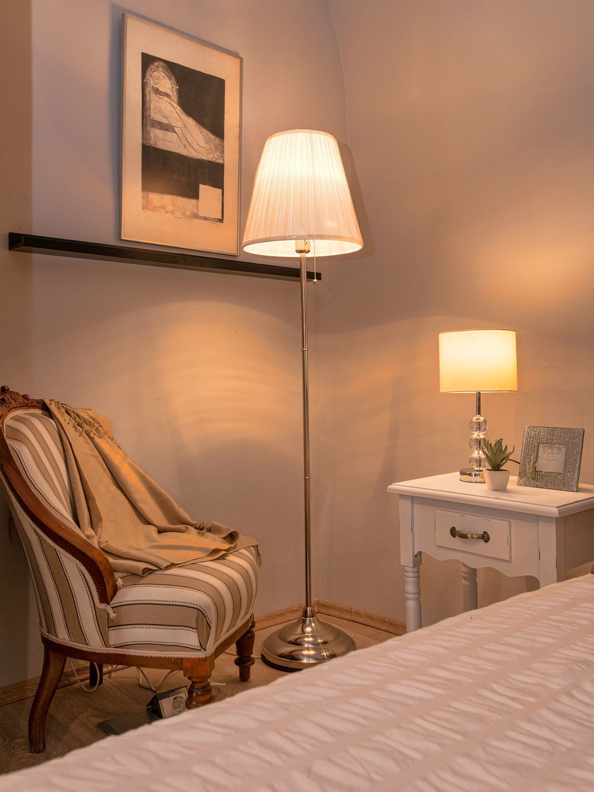 Un dormitorio con cama, una silla, mesita de noche con lámpara y un cuadro en la pared.