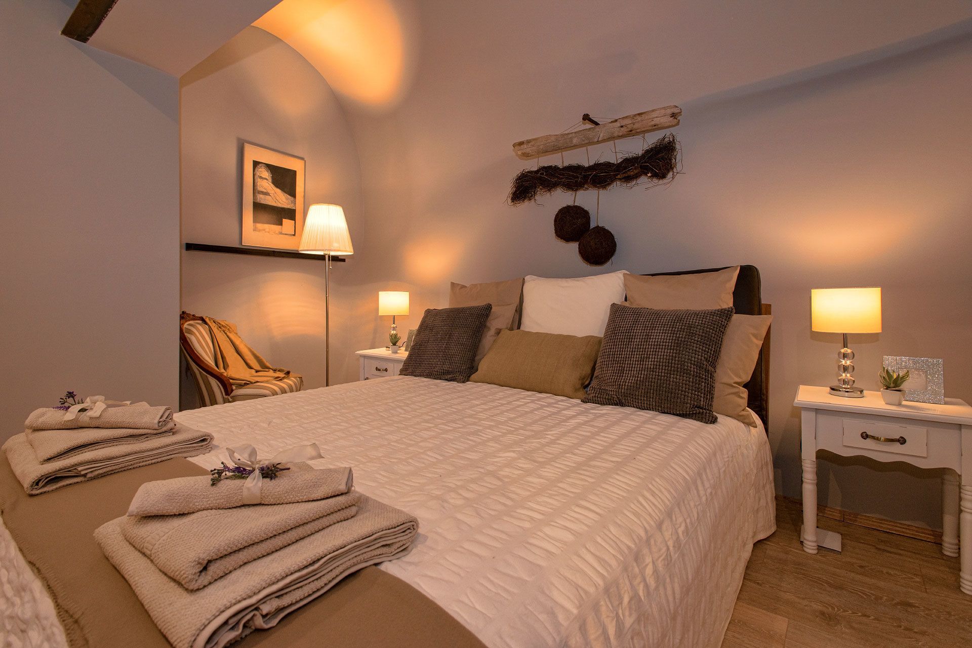 Ein ordentlich gemachtes Bett mit einer Bettdecke, zwei Nachttische mit Lampen, ein Sessel und ein an der Wand hängendes Kunstwerk.