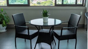 A room with a round table and four wicker chairs, a potted plant on the table, and large windows offering a view of a temple and cityscape. - Urbanite Hotel (Vientiane)