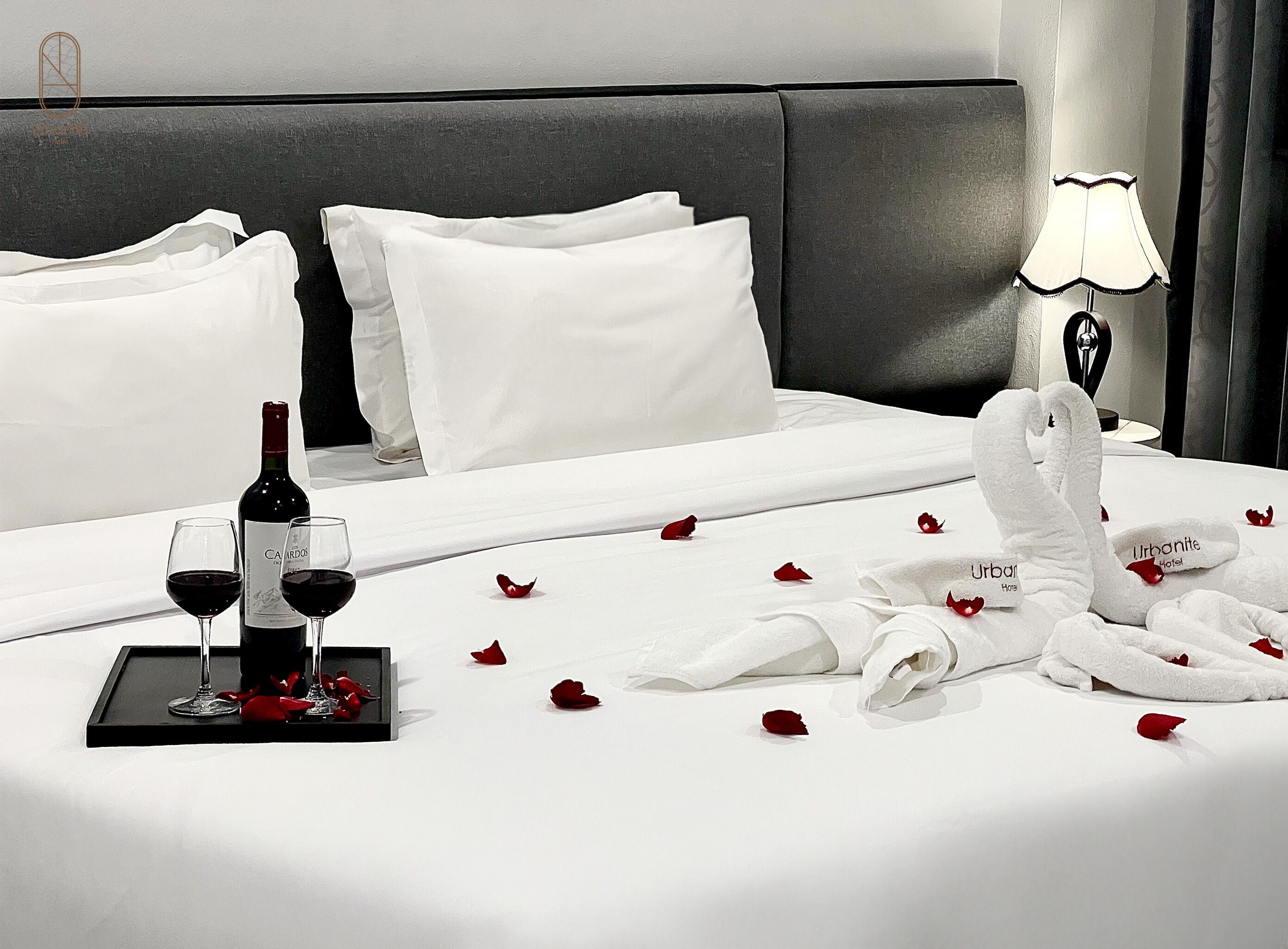 A hotel room with a neatly made bed, white linens, a bottle of wine, two glasses of wine, and rose petals scattered on the bed. - Urbanite Hotel (Vientiane)