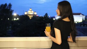 A person holding a glass of orange juice, standing on a balcony with a view of a building with illuminated domes in the background. - Urbanite Hotel (Vientiane)