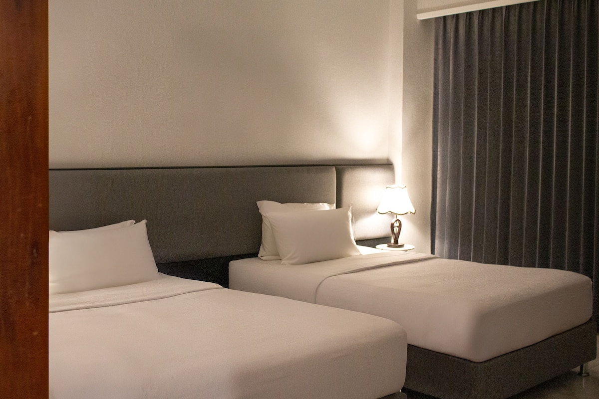 Ein Hotelzimmer mit zwei Betten, einem Kopfteil, einem Nachttisch mit Lampe und einem Fenster mit Vorhängen.