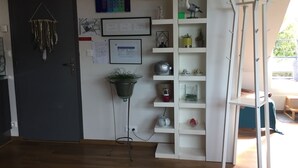 Huoneessa on tumma ovi, seinälle kiinnitetty hylly, jossa on erilaisia esineitä, roikkuva kasvi ja ikkuna, josta avautuu näkymä vehreään maisemaan.