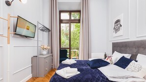 A modern bedroom with a large bed, a television mounted on the wall, a window with curtains, and a view of greenery outside. - The Old Town View (Kraków)