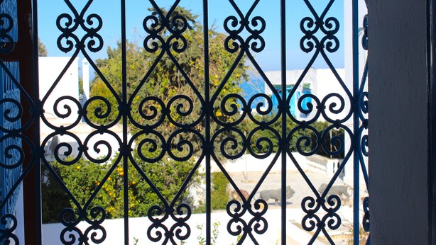 A wrought iron gate with intricate designs, leading to a building with white walls and a blue sky in the background. - Cosy Marsa Plage (La Marsa)