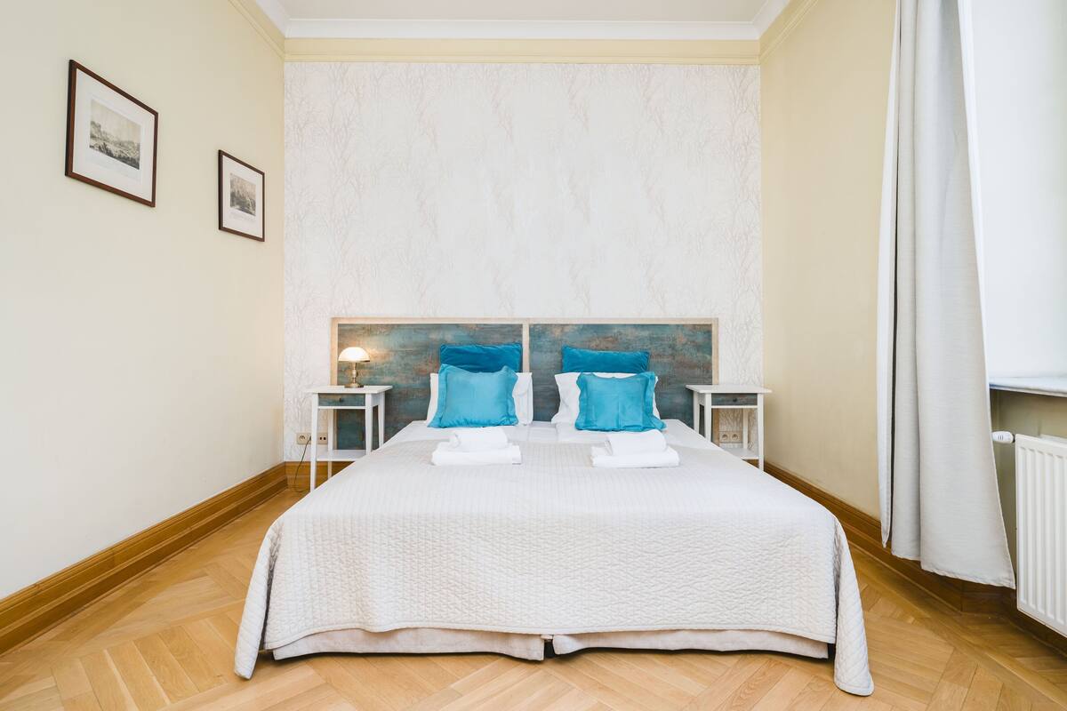 Een tweepersoonsbed met een witte geborduurde sprei en twee blauwe kussens, met nachtkastjes en lampen ernaast.