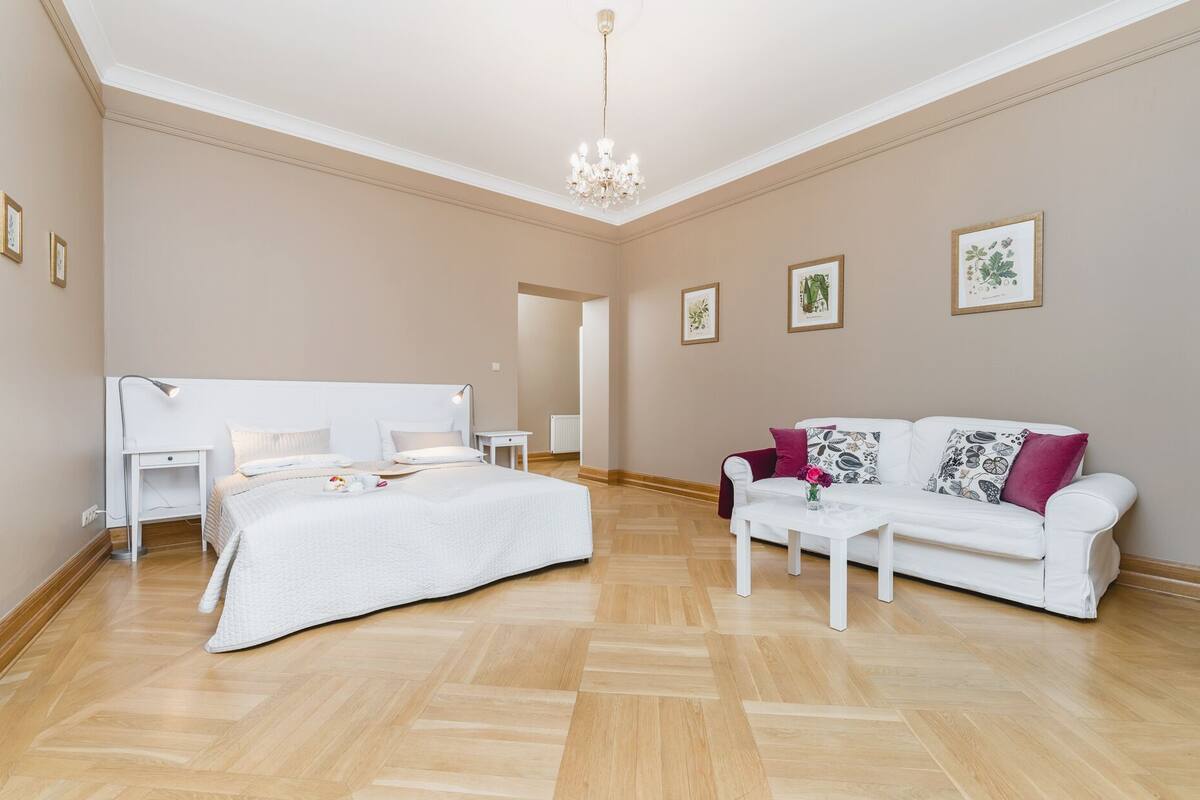 Een ruime kamer met een groot bed, een witte bank en een kleine tafel. Er hangen ingelijste foto's aan de muren en een kroonluchter aan het plafond.