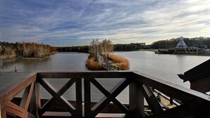 Een houten terras met uitzicht op een kalme rivier, bomen en een gebouw in de verte.