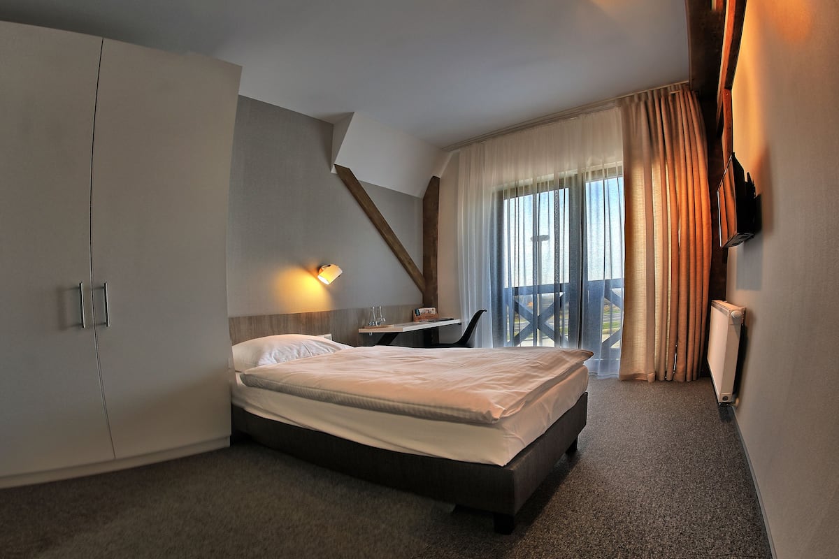 Habitación de hotel con cama, escritorio, silla, televisor y balcón con cortinas.