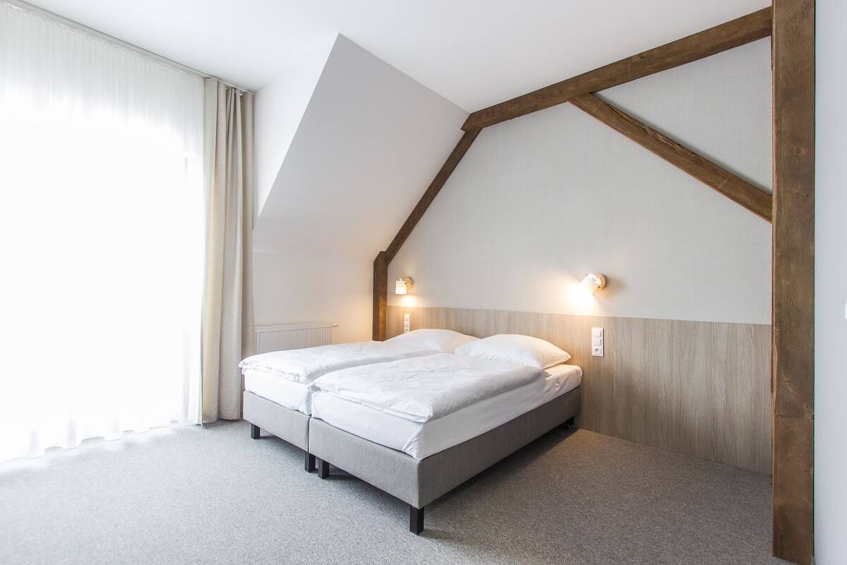Un dormitorio con techo inclinado, una cama doble, dos lámparas de noche y un ventanal con cortinas.