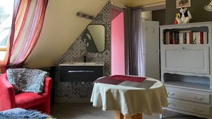 A compact room with a red chair, a small table with a white tablecloth, a vanity with a mirror, and a bookshelf.