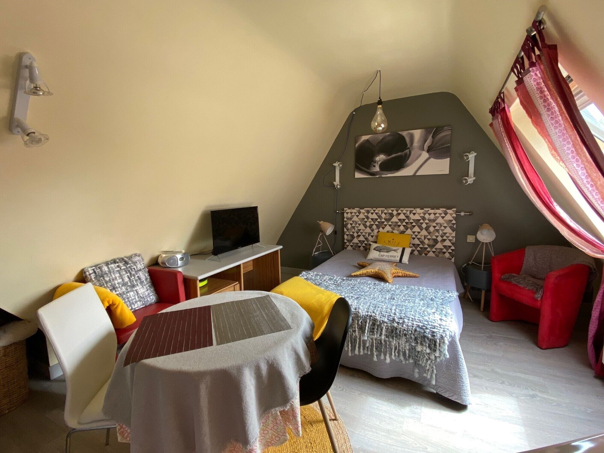 Een compacte slaapkamer met een bed, een bureau, een stoel, een televisie en een rode fauteuil.